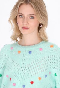 Maglione lavorato a maglia verde menta con un motivo testurizzato e motivi a cuore colorati in rosa, arancione, viola e rosso. Collo rotondo.