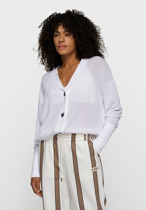 Femme aux cheveux bouclés portant un pull blanc ample boutonné et un pantalon blanc à rayures verticales marron avec des poches à cordon.