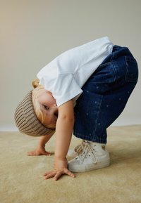 Camiseta blanca, pantalones vaqueros azules con lunares, gorro de punto beige y zapatillas blancas de caña alta; niño agachado apoyado en manos y rodillas.