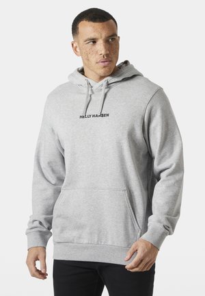 Hombre con pecas que lleva una sudadera con capucha Helly Hansen de color gris claro con bolsillo tipo canguro, mirando hacia un lado, de pie frente a un fondo liso.