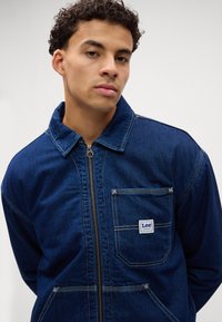 Donkerblauwe denimjack met een ritssluiting aan de voorkant, puntige kraag en borstzak met een geweven Lee-label. Zichtbare stiksels accentueren het geheel.
