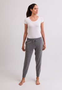 Graue Jogginghose mit einem Kordelzugbund, kombiniert mit einem figurbetonten weißen T-Shirt, barfuß auf einem neutralen Hintergrund stehend.