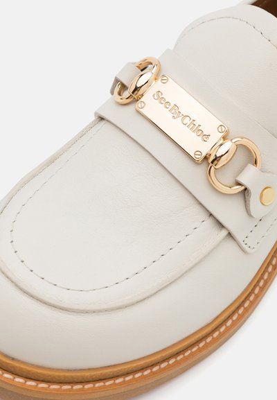 Loafers din piele bej cu detaliu din metal auriu, prezentând o placă rectangulară gravată cu "SeeByChloé" și talpă cusută în contrast.