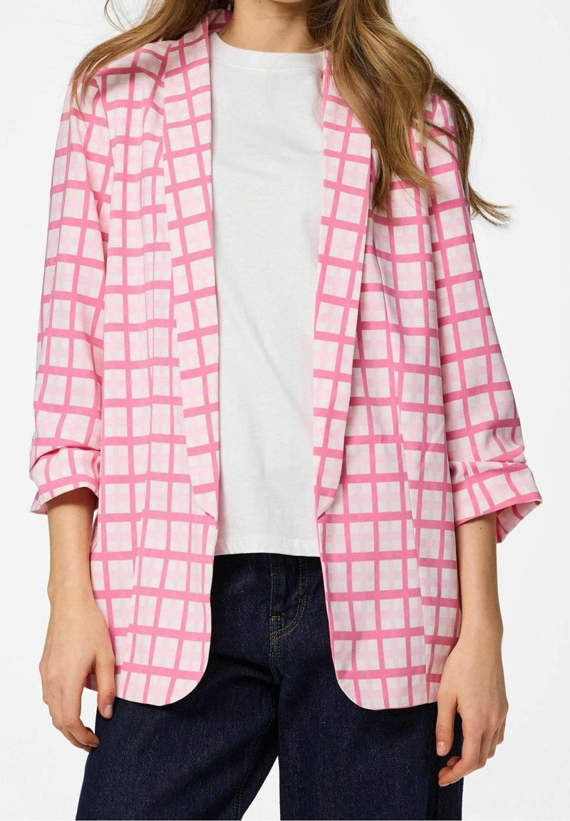Femme portant un blazer à carreaux rose et blanc sur une chemise blanche et un jean bleu foncé avec les manches retroussées.