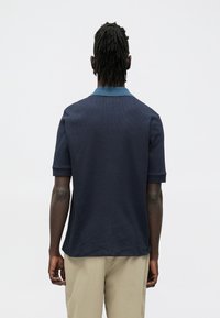 Tmavomodrá polokošeľa s textúrovanou tkaninou, krátkymi rukávmi a svetlejším modrým golierom. Má voľný strih a rovný lem.