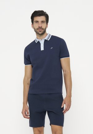 Mand iført en navy blå poloshirt med hvid krave og kant, kombineret med navy blå shorts, stående mod en ensfarvet baggrund.