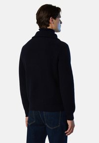 Marineblauer, gerippter Strickpullover mit hohem Kragen, taillierter Passform und langen Ärmeln, kombiniert mit blauen Denim-Jeans. Rückansicht dargestellt.