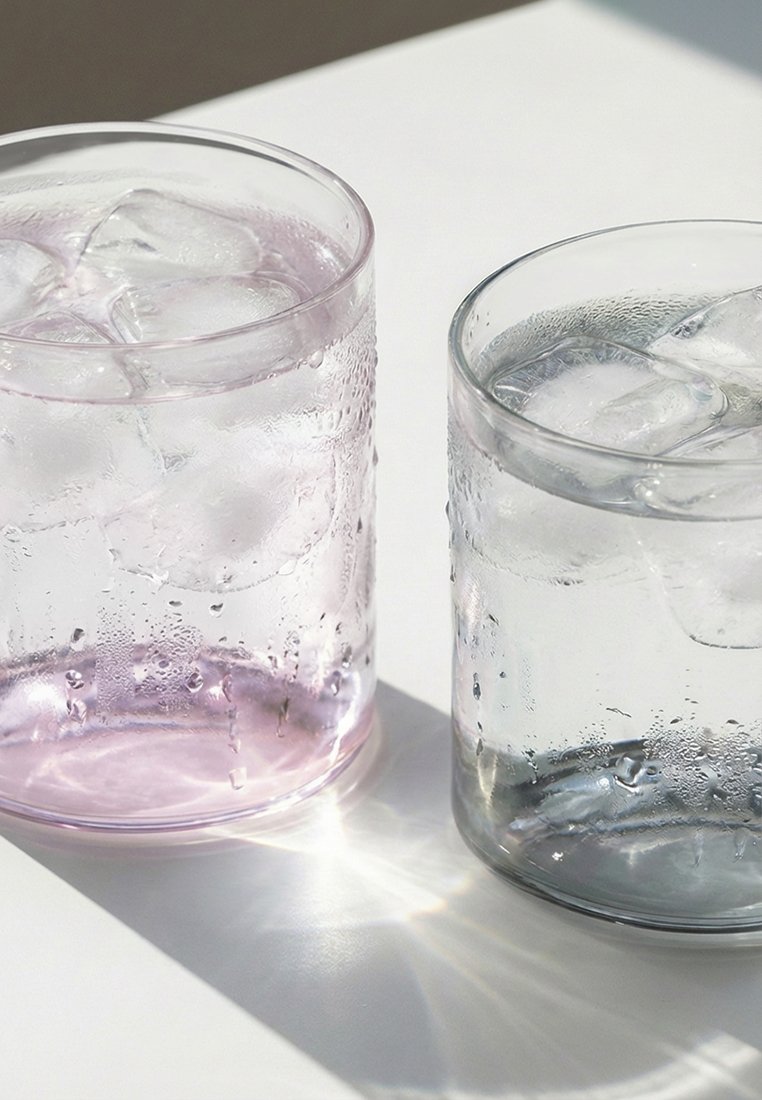 Dwa schłodzone przezroczyste szklanki z kostkami lodu, jedna z lekkim różowym odcieniem, druga przezroczysta, na białej powierzchni w świetle słonecznym.