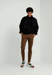 Young man standing with hands in pockets wearing black jacket, black turtleneck, brown pants, and multicolored sneakers on plain background.