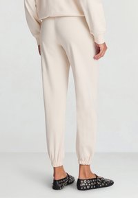 Joggers color crema con polsini elasticizzati, realizzati in tessuto morbido. Scarpe slip-on nere con dettagli di occhielli argento. Vista posteriore dell'outfit.