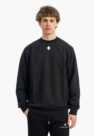 Jeune homme aux cheveux bouclés portant un sweat-shirt noir et un pantalon assorti avec un logo blanc en forme de pixel et le texte « COUNTY OF MILAN ».