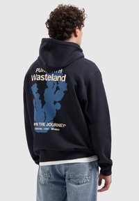 Man met krullend haar, gekleed in een marineblauwe hoodie met een blauw plantmotief en de tekst "wasteland" op de rug, in combinatie met lichtblauwe jeans.