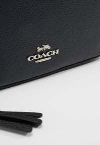 Černá kožená taška s texturovaným povrchem, vybavená stříbrným logem Coach a dekorativní třásní na boku.