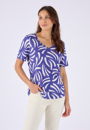 Donna con i capelli lunghi che indossa una maglietta a V blu e bianca con motivo astratto e pantaloni chiari, in piedi con una mano in tasca.