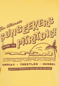 Kollane puuvillane t-särk, millel on pruun graafiline disain koos tekstiga ja joonistustega rannast, palmipuust ja päikesest, pealkirjaga "The Ultimate Sunseekers Paradise".