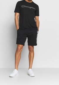 Man standing with hands in pockets wearing black Champion t-shirt, black shorts with logo on leg, and white sneakers against plain background.