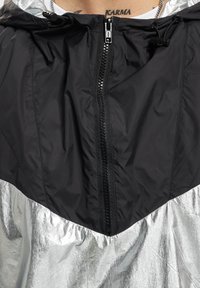 Schwarze und silberne Windjacke mit Reißverschluss, Kapuze und strukturiertem, metallischem Stoff. Verfügt über ein zweifarbiges Design und einen elastischen Saum.