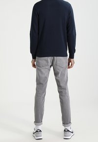 Felpa blu navy, jeans grigi slim-fit, sneaker bianche e nere. Tessuto liscio, orli risvoltati e dettagli delle tasche visibili.