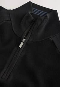 Zwarte pullover met rits en hoge kraag, met geribbelde textuur en een zilveren ritssluiting, afgewerkt met een blauwgestreepte binnenkant.