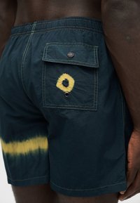 Short de bain bleu foncé avec des cercles jaune tie-dye, une poche rabat avec bouton, porté par une personne montrant la hanche et la main droite.