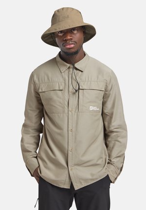 Homme portant une chemise kaki boutonnée avec poches poitrine et chapeau bob assorti, mains dans les poches du pantalon noir, fond neutre.