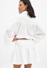 Blouse en lin blanc avec des manches volumineuses et un détail froncé au dos, associée à une jupe blanche évasée avec des plis horizontaux à la taille.