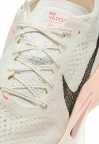 Buty Nike Vaporfly mają białą siateczkową cholewkę z czarnym logo Swoosh, różowymi akcentami, teksturowanymi sznurówkami oraz wyściełaną podeszwą.