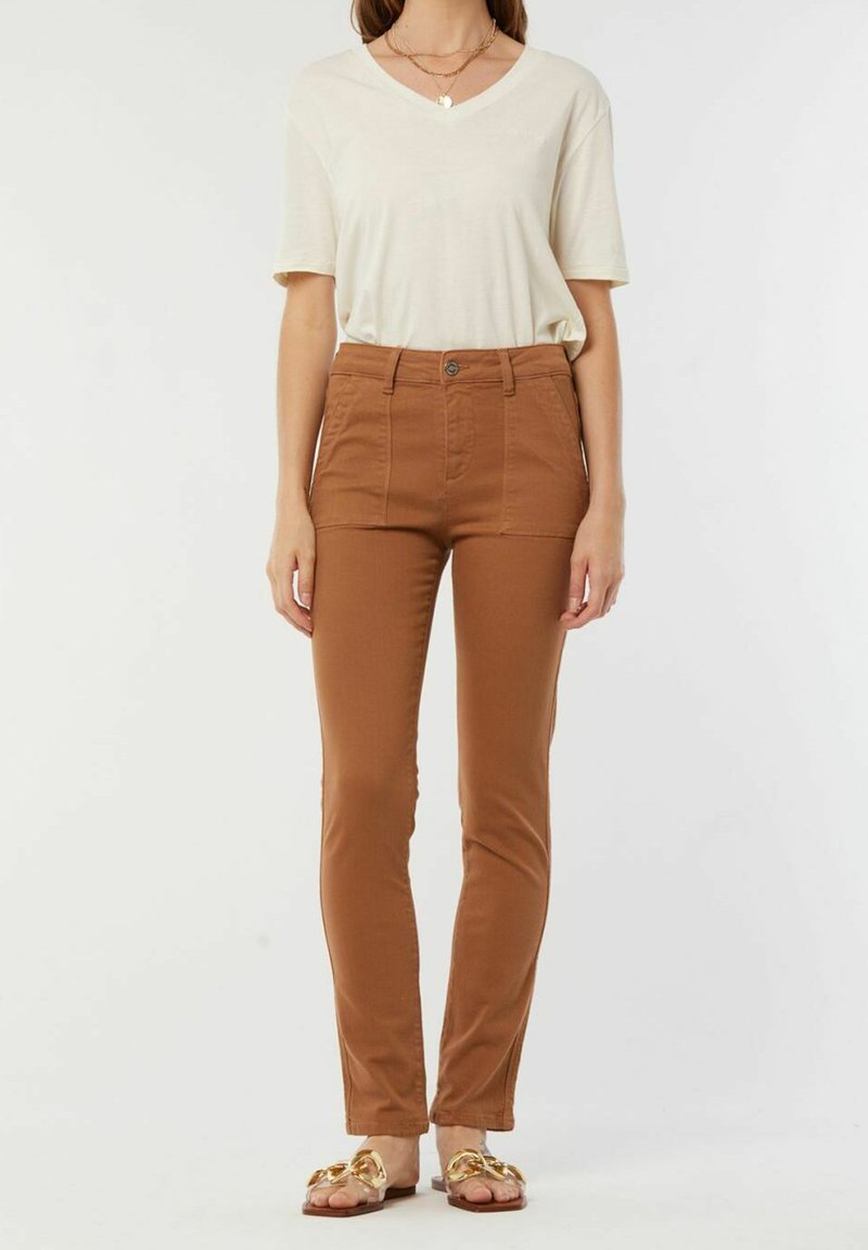T-shirt beige clair à col en V associé à un pantalon marron taille haute ajusté avec poches avant et fermeture par bouton. Sandales plates avec des accents dorés.