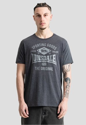 Giovane uomo con i capelli intrecciati che indossa una maglietta grigio scuro Lonsdale Sporting Goods, mostrando dei tatuaggi sull'avambraccio destro.