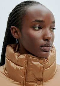 Braune gepolsterte Jacke mit hohem Kragen und Druckknöpfen. Glatte Textur und Glanz, mit einer Nahaufnahme eines Modells, das Gesicht und Hals zeigt.