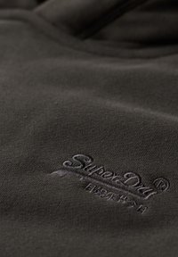 Tejido gris oscuro con una textura suave, que presenta el logotipo "SuperDry" bordado en un tono ligeramente más claro. Diseño suave y minimalista.