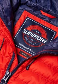 Czerwono-granatowa kurtka puchowa, wnętrze z etykietą "Superdry" i metką z rozmiarem "MAŁY". Materiał o fakturze z pikowanymi detalami.