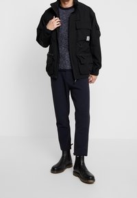 Giacca nera con zip e molte tasche, indossata sopra un maglione blu, abbinata a pantaloni scuri e stivaletti neri. Texture liscia.