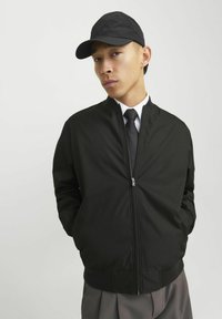 Chaqueta bomber negra con un cuello alto, cierre de cremallera y bolsillos laterales; combinada con una gorra negra a juego y una camisa blanca.
