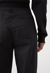 Personne portant un pantalon noir taille haute et un haut noir à manches longues, vue de dos avec une main détendue le long du corps.