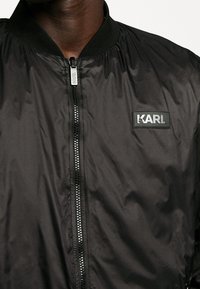 Czarna kurtka typu bomber z przednim zamkiem błyskawicznym, elastycznymi mankietami i prążkowanym kołnierzem. Na piersi znajduje się prostokątna czarna naszywka z logo „KARL”.
