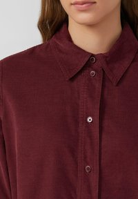 Chemise en velours côtelé bordeaux avec un col pointu, fermeture à boutons et des côtes verticales sur le tissu. Les boutons sont de la même couleur.