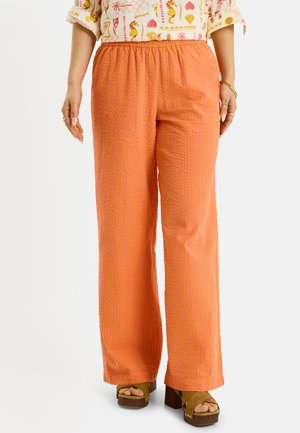 Mujer con pantalones anchos y texturizados naranjas, blusa estampada con mangas remangadas y sandalias de plataforma marrones, de pie con los brazos relajados.