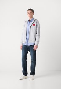 Lys grå hettegenser med glidelås og rødt logo, lagd over en blå og hvit stripete skjorte, sammen med mørke jeans og hvite joggesko.