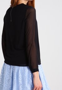 Donna che indossa un top nero trasparente a maniche lunghe con cerniera posteriore e una gonna di pizzo azzurro chiaro, in piedi contro uno sfondo bianco.