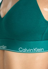 Cevna bombažna braletka z globokim V-izrezom. Beli logotip "Calvin Klein" na elastičnem traku spodaj. Gladka tekstura, preprost stil.