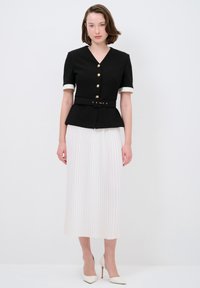 Schwarze Jacke mit kurzen Ärmeln, goldenen Knöpfen und tailliertem Design; kombiniert mit einem weißen, plissierten Midikleid und klassischen weißen High Heels.