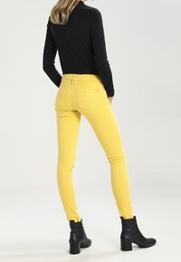 Persona con pantalones ajustados amarillos, blusa negra de manga larga y botas negras hasta el tobillo, de pie con las manos en los bolsillos y mirando hacia el lado opuesto.