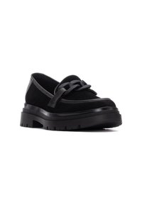 Zapato negro de ante tipo slip-on con una suela de plataforma gruesa, que presenta una correa de cuero decorativa sobre el empeine y detalles de costura en contraste.