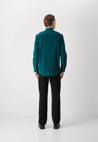 Camisa de pana color verde azulado con cuello tipo polo y mangas largas, combinada con pantalones negros. Presenta un dobladillo redondeado y detalles en la parte posterior.