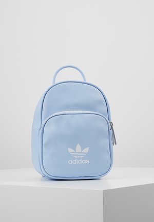 Lyseblå Adidas mini-rygsæk med lynlåslomme foran og logo, placeret på en hvid piedestal mod en ensfarvet lysegrå baggrund.