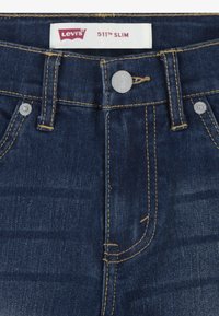 Levi's 511 Slim džinsi tumši zilā denimā ar dzeltenām šuvēm. Iespējas: pogas aizdare, priekšējās kabatas un jostas birka.