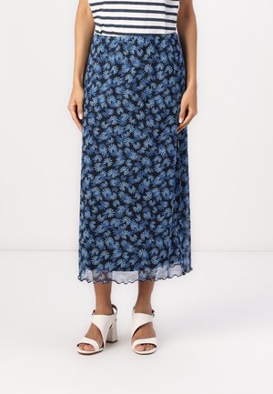Donna che indossa una gonna trasparente blu navy con motivo floreale azzurro chiaro, abbinata a sandali bianchi con tacco e una maglia a righe.