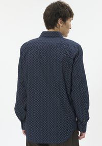Mann, der sich abwendet, trägt ein langärmliges navyblaues Hemd mit einem kleinen Rautenmuster auf weißem Hintergrund.
