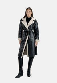 Cappotto in pelle nera con un rivestimento in pelliccia crema, ampio colletto, cintura in vita e due tasche frontali. Indossato sopra un top nero e stivali di pelle al ginocchio.